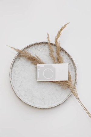 Photo for Blank textured paper business card mockup. Ceramic plate isolated on white table background. Boho stationery with dry grass. Branding template. Minimal composition, vertical flat lay, top view. - Royalty Free Image