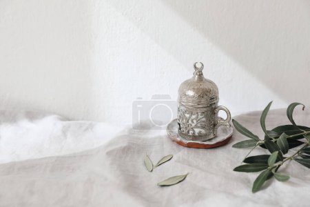 Photo for Decorative silver Turkish cup of tea. Green olive tree branches on white linen table cloth in sunlight. Festive still life for Muslim community holy month Ramadan Kareem, blurred background, shadows. - Royalty Free Image