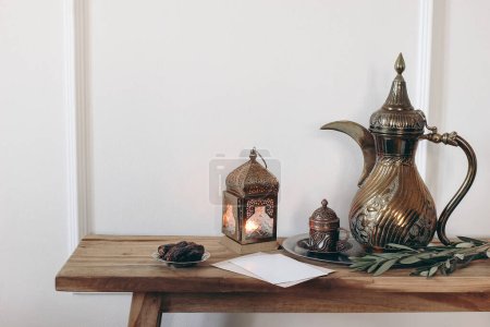 Ramadan Kareem still life. Ornamental burning lantern, cup and golden arabic dallah coffee pot. Green olive tree branches and date fruit on old wooden table. White wallbackground, muslim Iftar dinner.