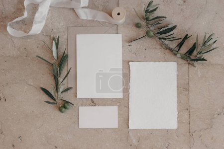 Photo for Italian summer wedding stationery set. Mock-up scene with blank paper greeting, RSVP cards on beige marble tiles background. Green olive tree branches, silk ribbonMediterranean flat lay, top view. - Royalty Free Image