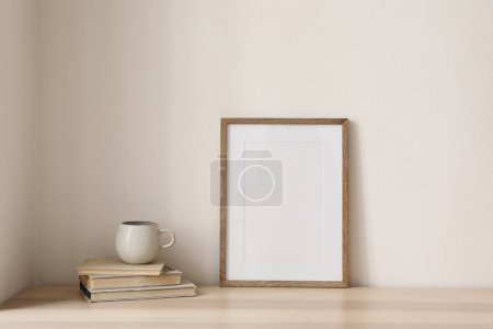 Photo for Empty wooden picture frame, poster mockup on wooden table. Cup of tea, coffee on old books. Breakfast concept. Beige wall background. Working space, home office, modern art display, elegant interior. - Royalty Free Image