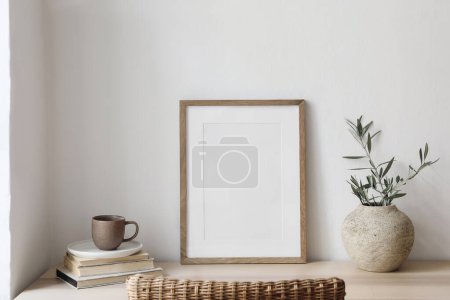 Photo for Closeup of picture frame, poster mockup. Vase with olive tree branches on wooden table. Blurred rattan chair. Cup of coffee, books. Summer artistic Mediterranean interior, working space, home office. - Royalty Free Image