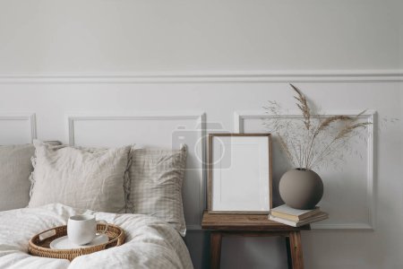 Photo for Elegant boho bedroom interior. Vertical wooden picture frame mockup on old bedside table. Cup of coffee. Ball vase with dry grass. Beige linen throw bedding. White wall background, Scandinavian home. - Royalty Free Image