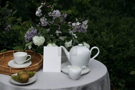 Nahaufnahme einer leeren Grußkarte, einer Hochzeitseinladung zum Geburtstag. Teeparty im grünen Garten. Teekanne, Milchkanne, Birnenobst auf dem Tisch mit Leinen-Tischdecke, Tasse Kaffee. Flieder, Viburnum-Blumen