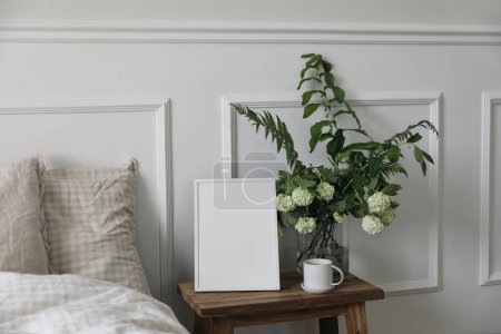 Photo for White picture frame mockup and cup of coffee on wooden night stand. White viburnum, fern and solomons seal flowers bouquet. Bedroom view, beige pillows, linen blanket in bed. Home interior decor. - Royalty Free Image