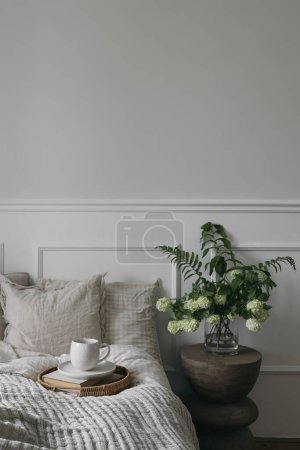 Breakfast in bed. Cup of coffee, wicker tray. Bouquet of white viburnum, fern and solomons seal flowers. Modern boho night stand. Bedroom view, beige pillows, blanket. Blurred background, vertical.