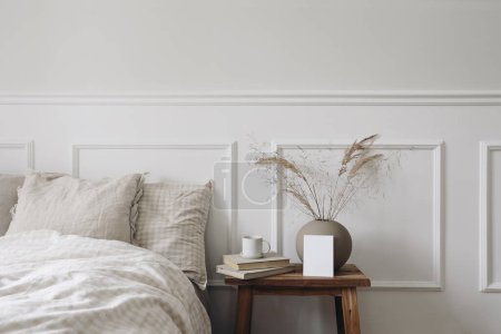 Photo for Elegant Scandinavian bedroom interior. Blank greeting card, invitation mockup on bedside table. Cup of coffee, old books. Ball vase with dry grass. Beige linen throw bedding, white wall background. - Royalty Free Image