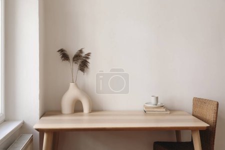 Photo for Wooden table, desk near window. Modern organic shaped vase with dry flowers, grass. Cup of tea, coffee, old books. Rattan chair at home. Minimal Scandi boho interior, beige wall background mockup. - Royalty Free Image