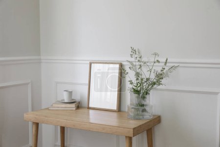 Photo for Minimal working space, home office. Elegant corner still life. Cup of coffee, books. Empty vertical picture frame mockup. Wooden desk, table, vase, green grasses and cow parsley. Scandi interior, side - Royalty Free Image