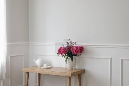 Photo for Living room still life. Vase with pink peonies flowers. Cup of tea, coffee and tea pot on wooden table near window with curtains. Romantic breakfast, empty white wall background with elegant moulding. - Royalty Free Image