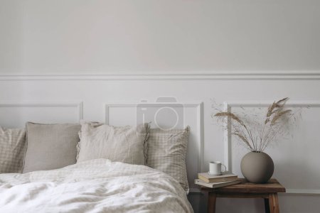 Photo for Moody boho bedroom interior. Vintage wooden bedside table. Round modern vase with dry grass. Cup of coffee on old books. Beige linen throw bedding. White wall background, Scandinavian home, no people. - Royalty Free Image