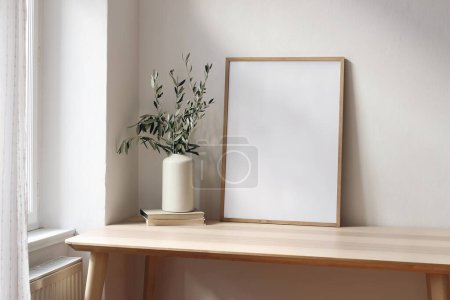 Blank wooden vertical picture frame mockup, books on office table near window in sunlight. Mediterranean interior. Elegant vase with olive tree branches. Living room, modern summer design.