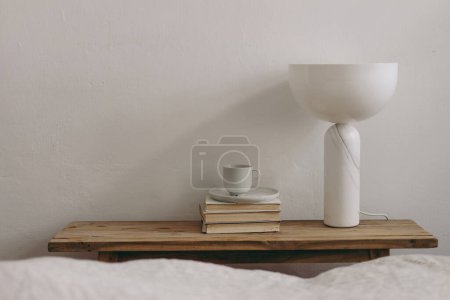 Photo for Elegant home, interior. Cup of coffee, books and modern marble geometric lamp on vintage teak wooden bench, table. Empty white wall mockup background, blurred bed. Scandinavian interior, bedroom. - Royalty Free Image