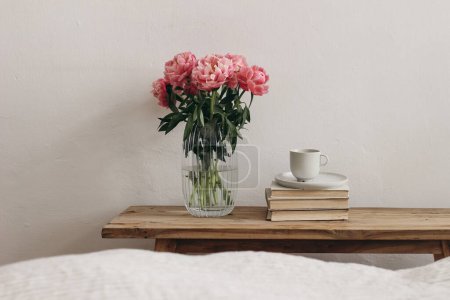 Photo for Feminine interior, still life. Rippled glass vase with coral pink peony flowers. Floral arrangement. Cup of coffee, books on vintage teak wooden bench, table. Empty white wall background. Blurred bed - Royalty Free Image