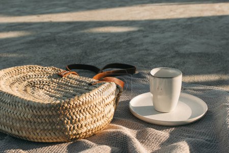 Photo for Elegant wicker straw bag on beige towel in sunlight. Cup of coffee, drink and plate. Concrete ground, sand beach. Resting at the resort, collection of beach items. Traveling, vacation, relaxation - Royalty Free Image