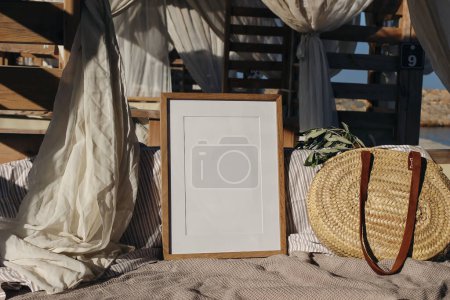Photo for Blank picture frame, poster mockup in beach lounger, gazebo with canopy in resort at sea. Wicker straw bag with green olive tree branches in sunlight, summer vacation, traveling concept. - Royalty Free Image