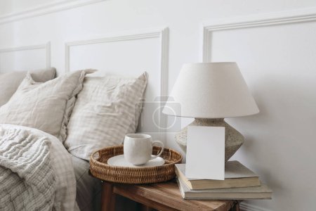 Photo for Closeup of blank greeting card, invitation mockup. Breakfast in bed concept. Cup of coffee, table lamp with linen shade, vintage wooden night stand. Checkered beige pillows. Blurred background. - Royalty Free Image