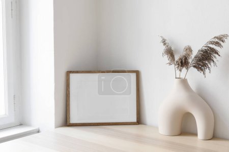 Photo for Dry grass, reed plant bouquet in modern ceramic vase. Empty photo frame mockup against white wall on wooden table near window. Minimalistic Scandinavian style, elegant boho interior. Living room. - Royalty Free Image