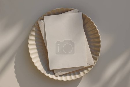 Photo for Summer greeting card mockup. Blank wedding invitation, template with craft envelope on beige ceramic scallop dinner plate in sunlight. Beige table background. Minimal composition, flatlay, top view. - Royalty Free Image
