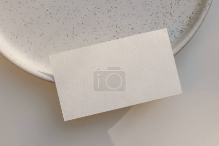 Photo for Summer branding stationery. Closeup of textured blank business card, invitation mockup on dotted, speckled ceramic plate in sunlight. Beige table background. Elegant flat lay, top view, no people. - Royalty Free Image