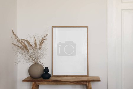 Photo for Vertical wooden frame mockup for artwork, photo, print and painting presentation. Black and beige vases with dry grass on wooden bench table. Elegant boho living room interior, white wall, doors. - Royalty Free Image