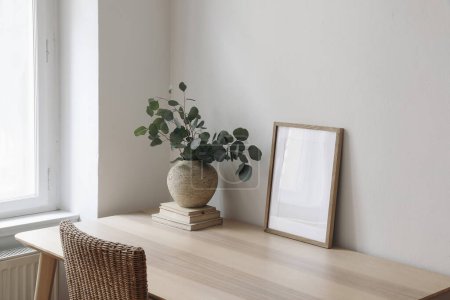 Photo for Stylish interior. Living room with poster frame mock up on wooden table, desk. Eucalyptus tree branches, rattan chair near window. Minimalist concept of home decor. Scandinavian design, blank template - Royalty Free Image