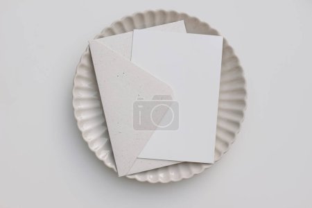 Photo for Summer neutral wedding stationery. Bank greeting card mockup with beige envelope. Invitation, template on ceramic scallop dinner plate. White table background. Elegant composition, flatlay, top view. - Royalty Free Image