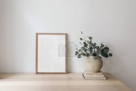 Photo for Scandinavian living room interior with poster frame mock up on wooden table, desk. Textured vase with eucalyptus tree branches, pile of old books, white wall. Minimalist concept of home office decor. - Royalty Free Image