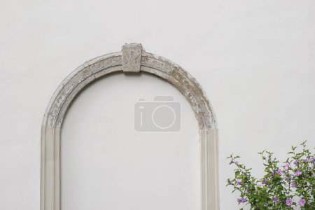 Decorative arch and semi vault above niche with stone pillars. Old walled gate, entrance. Architectural detail of historical buildingin. White wall background. Hibiscus bush, elegant masonry facade