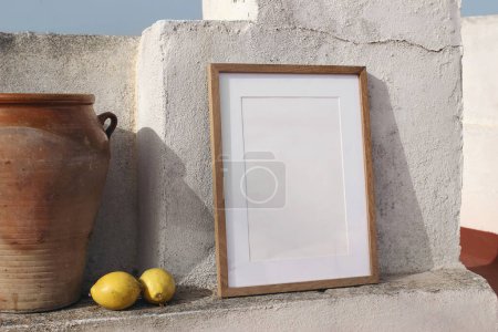 Photo for Blank vertical wooden frame picture mock up with vintage olive clay storage pot, vase. White old textured white wall in sunlight. Lateral view, no people, summer display background for art, posters. - Royalty Free Image