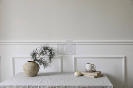 Photo for Elegant Christmas interior. Winter dinner table setting. Cup of coffee, old books and little white pumpkin. Pine tree branches in vintage vase. White wall background, classic moulding trim, no people - Royalty Free Image