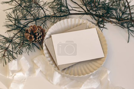 Photo for Christmas still life. Blank horizontal greeting card, invitation mockup on scalloped plate in sunlight. Pine cone, pine tree branches. White linen tablecloth, silk ribbon, winter gift concept. Flatlay - Royalty Free Image