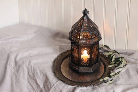 Photo for Burning black Moroccan, Arabic lantern with silver tray, olive tree twigs on table with linen tablecloth. White wooden wall background. Muslim holiday Ramadan Kareem home still life. Iftar dinner. - Royalty Free Image