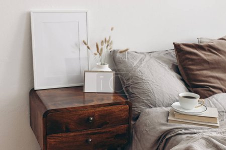 Photo for Elegant bedroom. Blank greeting card, invitation and picture frame mockup on old books. Brown, grey linen and velvet pillows. Wooden night stand with ceramic vase, dry grass, cup of coffee - Royalty Free Image