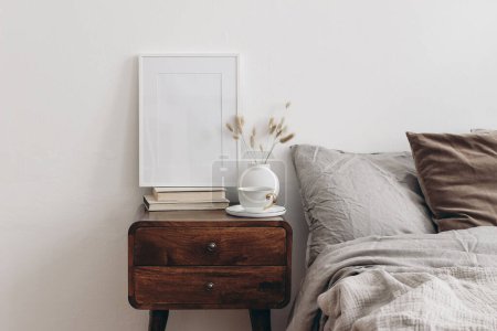 Photo for Vertical white picture frame mockup on old books. Elegant bedroom view. Brown, grey linen and velvet pillows, blanket. Wooden night stand with ceramic vase, dry grass and cup of coffee, Scandinavian - Royalty Free Image