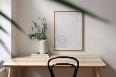 Photo for Empty vertical picture frame mockup. Wooden desk, table. Vase with olive branches, magazines. Elegant working space, home office. Summer interior design. Beige walls, blurred palm leaf overlay. - Royalty Free Image