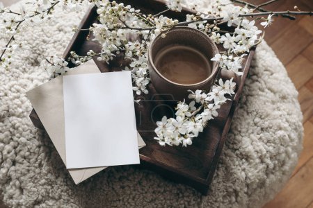 Spring still life composition. Greeting card mockup, cup of coffee. Feminine styled photo. Floral scene with blurred white cherry tree blossoms on wool taburet, stool. Wooden parquet floor, selective