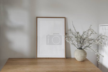 Photo for Vertical wooden picture frame, poster mockup in sunlight. Spring composition. Elegant interior, home office still life. Artistic display. Blooming cherry plum tree branches in vase, wooden table, desk - Royalty Free Image