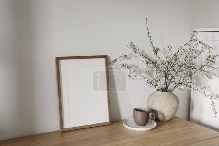 Photo for Vertical wooden picture frame, poster mockup in sunlight. Spring, easter composition. Eleganrnterior, home office still life. Blooming cherry plum tree branches in vase. Wooden table, lateral view. - Royalty Free Image