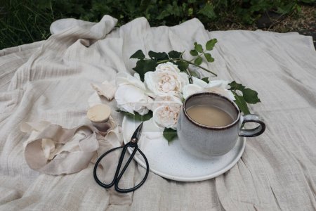 Photo for Elegant summer picnic in the garden still life. Pink nude colored roses, silk ribbons and vintage scissors on beige linen tablecloth. Cup of coffee. Blurred green grass background. Selective focus. - Royalty Free Image