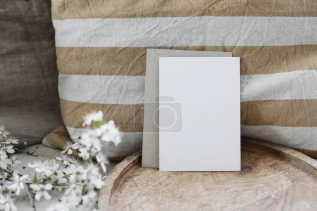 Photo for Spring still life composition. Blank greeting card mockup, craft envelope. Feminine styled photo. Floral scene. Blurred white cherry tree blossoms on wooden tray. Yellow striped linen cushion, front - Royalty Free Image