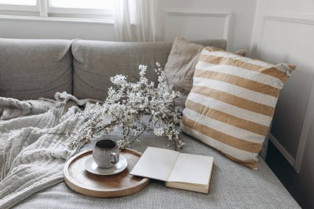 Photo for Modern spring scandinavian living room interior. Sofa with linen yellow striped cushions and cup of coffee. Cherry plum blossoms in vase. Open blank book, diary or notepad, elegant home office decor. - Royalty Free Image