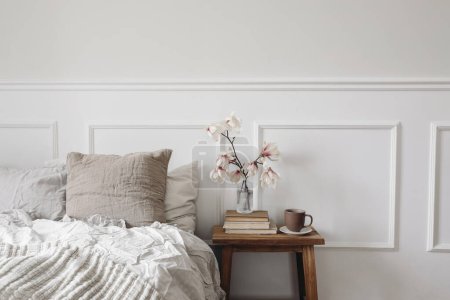 Photo for Elegant bedroom. Wooden night stand with fluted glass vase. Boolming magnolia tree branches. Cup of coffee on old books. Scandinavian interior. Linen bedding, white wall background, stucco decor. - Royalty Free Image