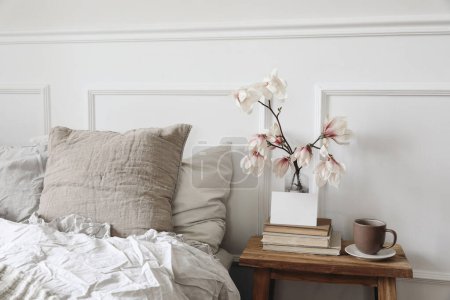 Photo for Blank greeting card, invitation mockup on old books. Elegant bedroom. Linen pillows, blanket. Wooden night stand, rippled glass vase. Blooming magnolia branches. Cup of coffee. Scandinavian interior. - Royalty Free Image