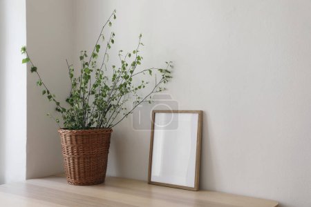 Photo for Spring, Easter still life. Elegant Scandinavian living room, home office. Empty vertical wooden picture frame mockup on desk, table. Wicker willow basket with green birch tree branches, side view. - Royalty Free Image