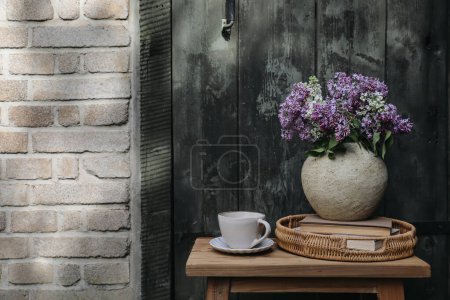Photo for Farm spring breakfast still life. Purple, white lilac flowers bouquet in textured vase with cup of coffee. Wicker tray, books. Blurred old wooden door background, shabby brick wall in sunlight. - Royalty Free Image