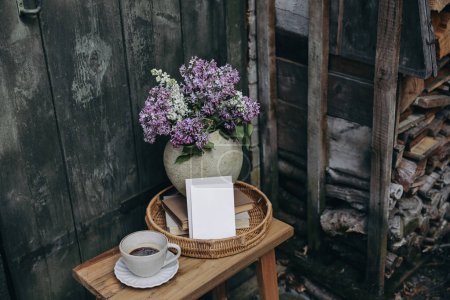Photo for Moody rustic spring outdoor still life. Purple, white lilac flowers bouquet in textured vase. Cup of coffee, books. Greeting card, invitation mockup, blurred old wooden door background. Pile of - Royalty Free Image