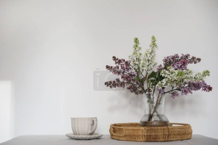 Photo for Moment of tranquility with cup of coffee, wicker tray. Floral bouquet of blooming purple and white lilacs branches in glass vase. Springtime breakfast scene, table, white wall background, selective - Royalty Free Image