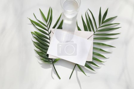 Photo for Blank paper, greeting card. Invitation mockup with fresh green palm leaves. Drinking lass with water in sunlight. Shadows on white table background. Flat lay, top view, aesthetic business brand - Royalty Free Image