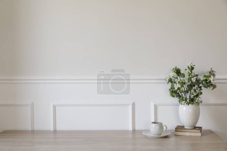 Ceramic vase with blooming apple tree branches. Cup of coffee, tea on wooden table, desk with old books. Scandi home interior. Spring breakfast still life. White wall mockup, stucco decor, copy space.